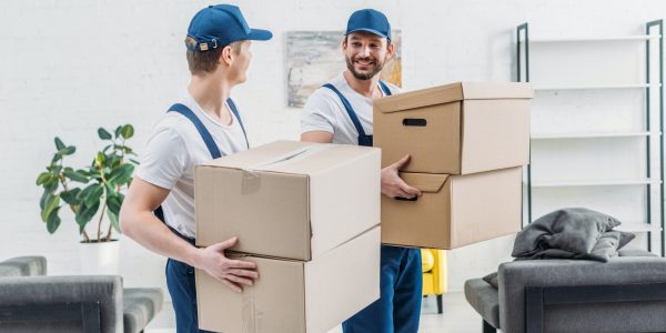 two-movers-in-uniform-looking-at-each-other-while-carrying-cardboard-boxes-in-apartment.jpg